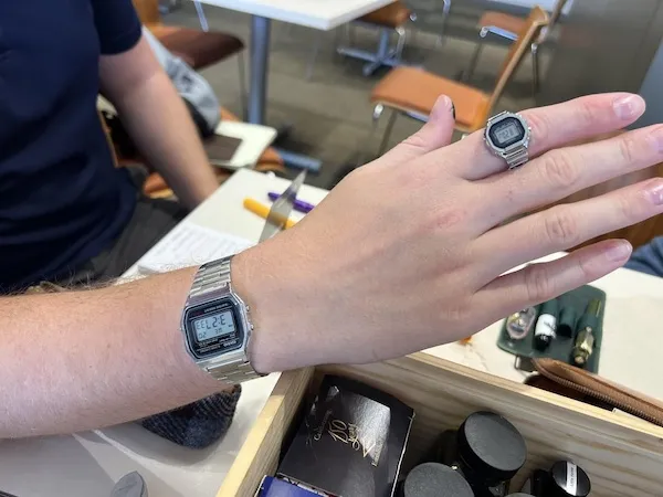 Ruka s digitálnymi hodinkami a prsteňom v tvare hodiniek na pracovnom stole v kaviarni.