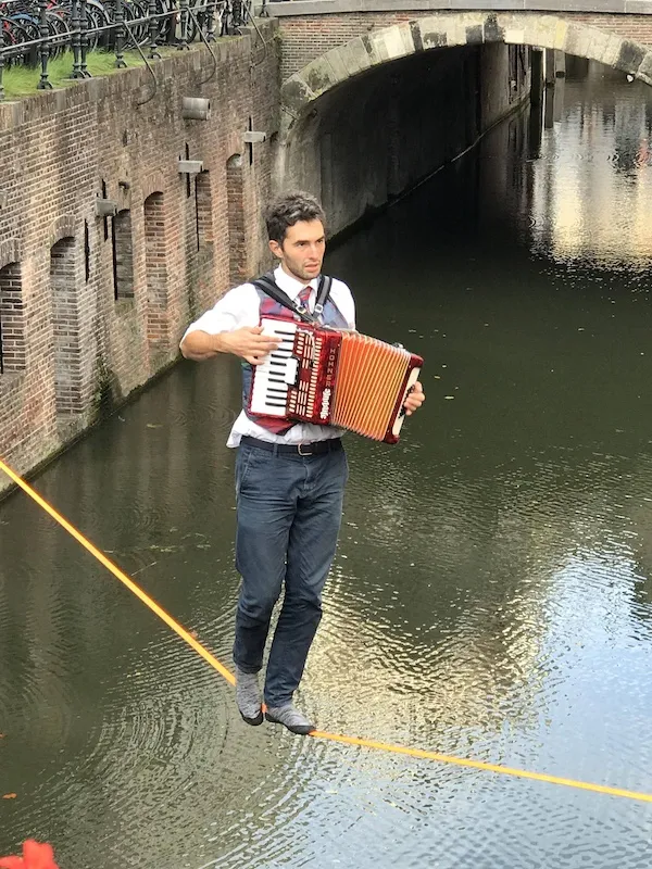 Muž hrá na akordeóne na lane nad vodou, v pozadí tehlový most a kanál.