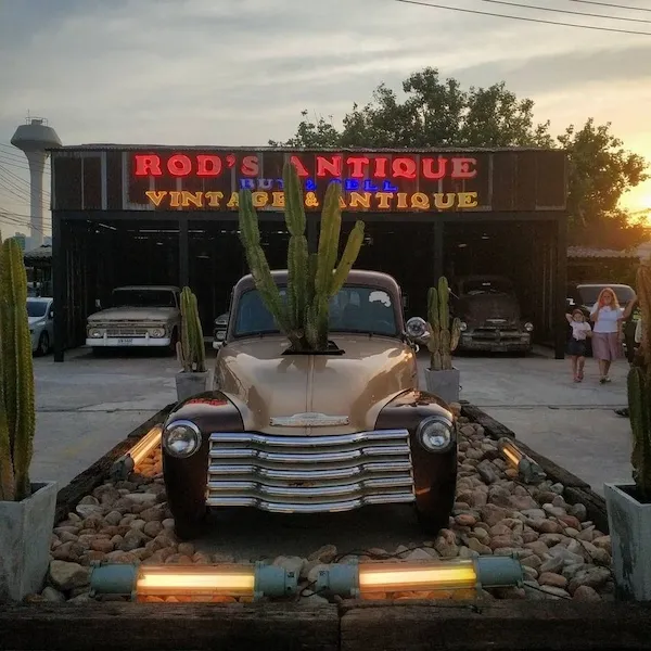 Staré retro auto pred obchodom s nápisom Rod's Antique, ozdobené kaktusmi, pri západe slnka.