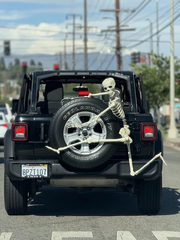 Kostra sedí na nárazníku terénneho auta, drží sa rezervného kolesa, uprostred cestnej premávky.