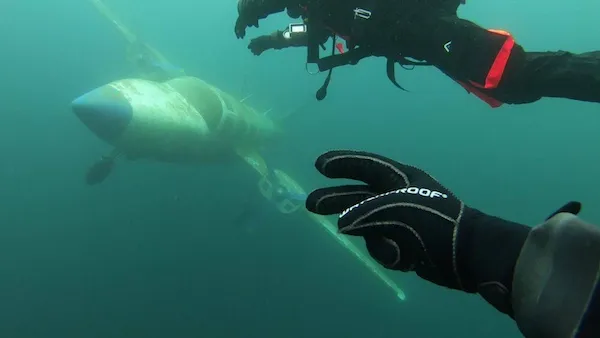Potápač skúma potopené lietadlo pod vodou, pričom detail rukavice je v popredí.