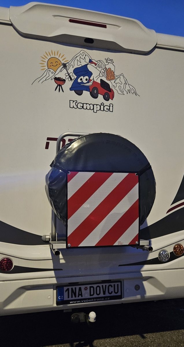 Karavan s nálepkou Kempici, hory v pozadí, úsmevné slnko, gril a pivo. ŠPZ: 1NA DOVCU.