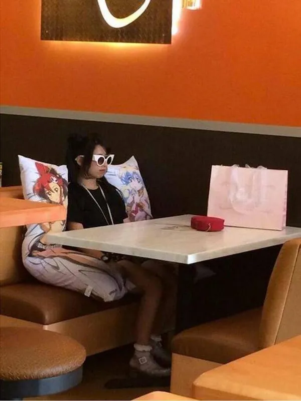 Osoba v reštaurácii sedí vedľa vankúšov s anime postavami, nosí slnečné okuliare a čierne oblečenie.