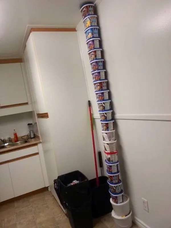 Veža z plastových kelímkov na maslo v kuchyni pri odpadkovom koši, dosahujúca až po strop.