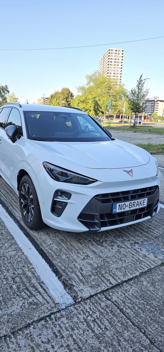 Biele auto parkujúce na ulici so zaujímavou poznávacou značkou NO BRAKE. Moderná architektúra v pozadí.