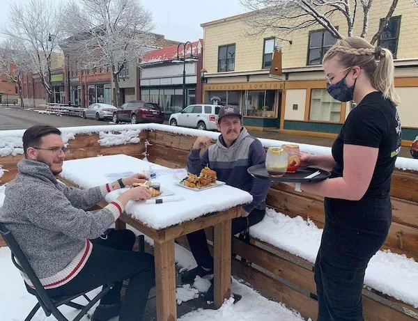 Žena obsluhuje dvoch mužov sediacich pri zasneženom stole na terase reštaurácie v zimnom meste.