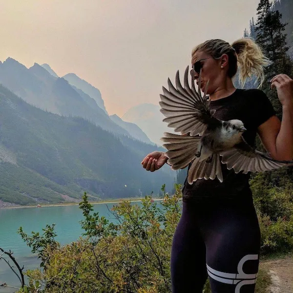 Žena pri jazere s vtákom v rukách, obklopená horskou scenériou.