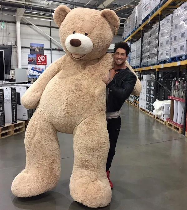 Obrovský plyšový medveď v sklade, usmievavý človek ho objíma, stojaci vedľa regálov plných tovaru.
