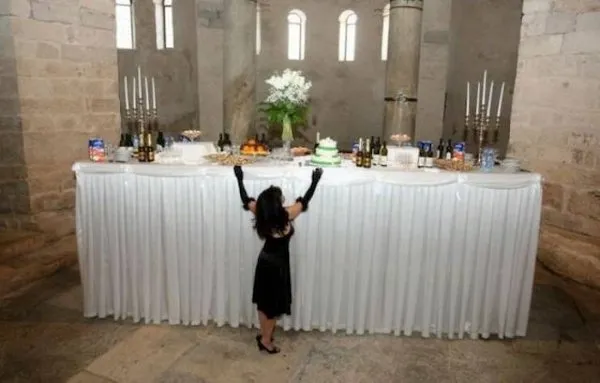Žena v čiernych šatách sa pokúša dosiahnuť na vysoko položený stôl so svadobnými pochúťkami v historickom priestore.