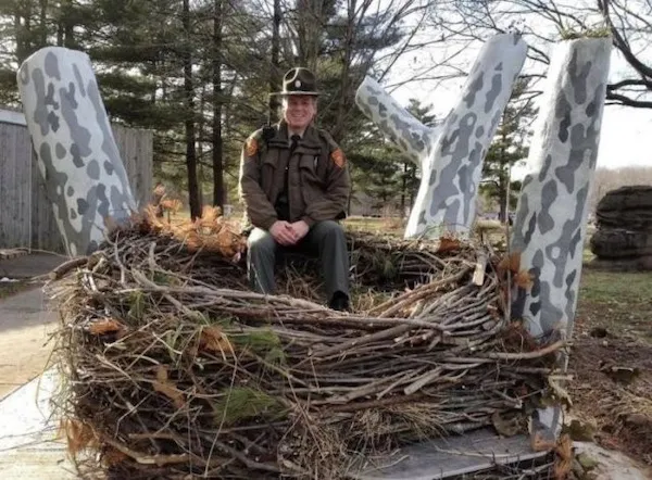Osoba v uniforme sedí v obrovskom hniezde z vetvičiek na vonkajšom pozadí stromov.