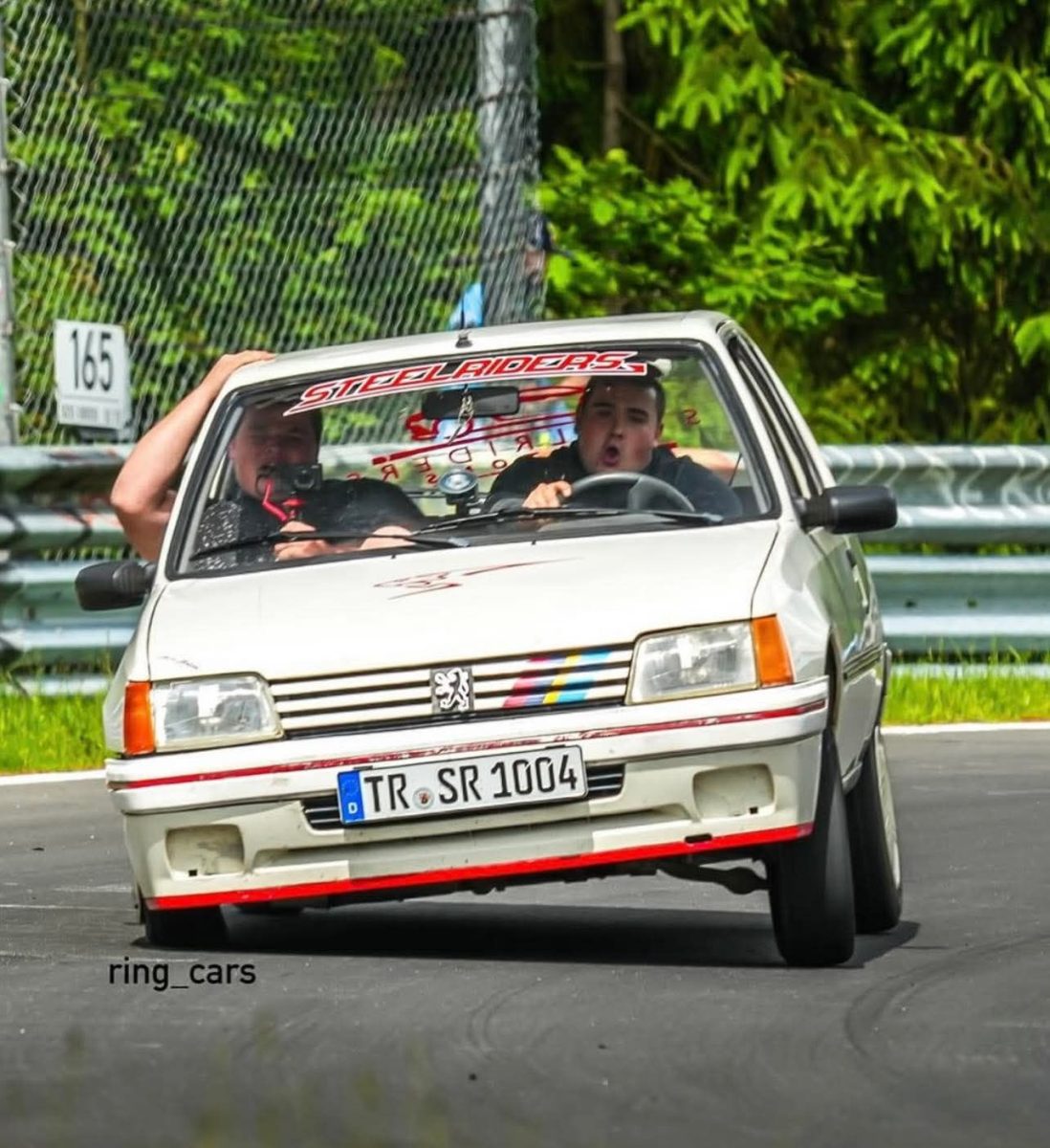 Auto Peugeot 205 v ostrej zákrute na pretekoch, adrenalínový moment na trati, dvaja pasažieri v interiéri.