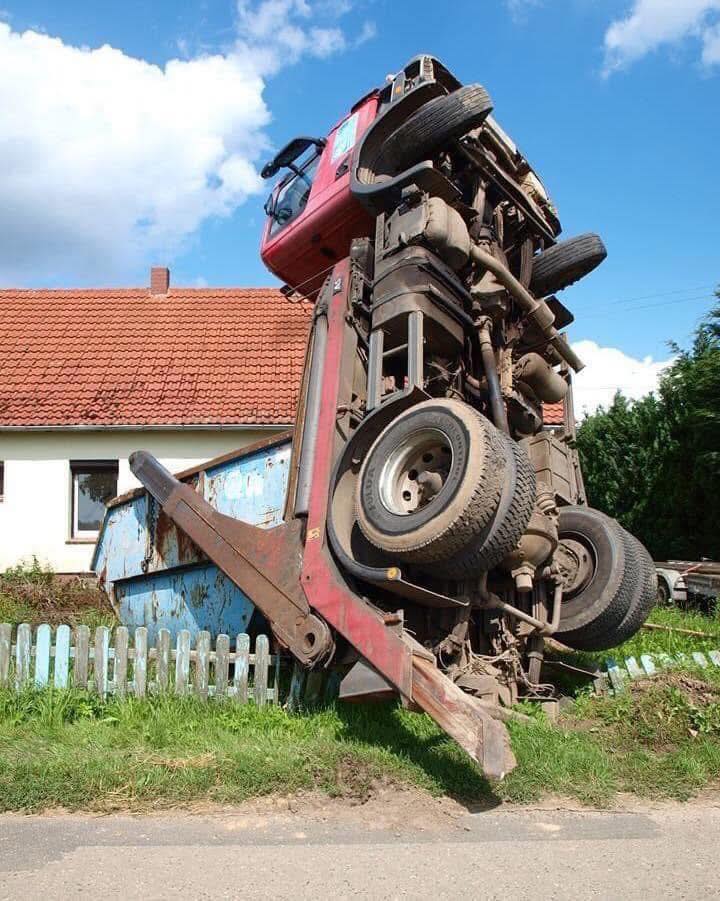 Nákladné auto zapadnuté v jame vedľa domu, zdvihnuté vertikálne, kolesá smerujúce nahor.