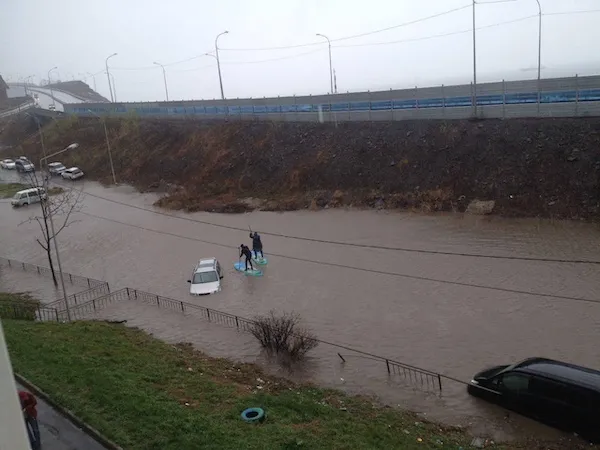 Povodeň v meste, autá uviaznuté vo vode, dvaja ľudia na surfe na zaplavenej ceste pod mostom.