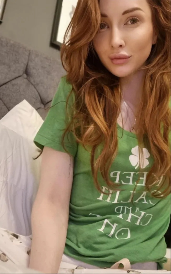 Osoba s dlhými ryšavými vlasmi v zelenej tričku sedí na posteli, pózuje pre selfie fotku.