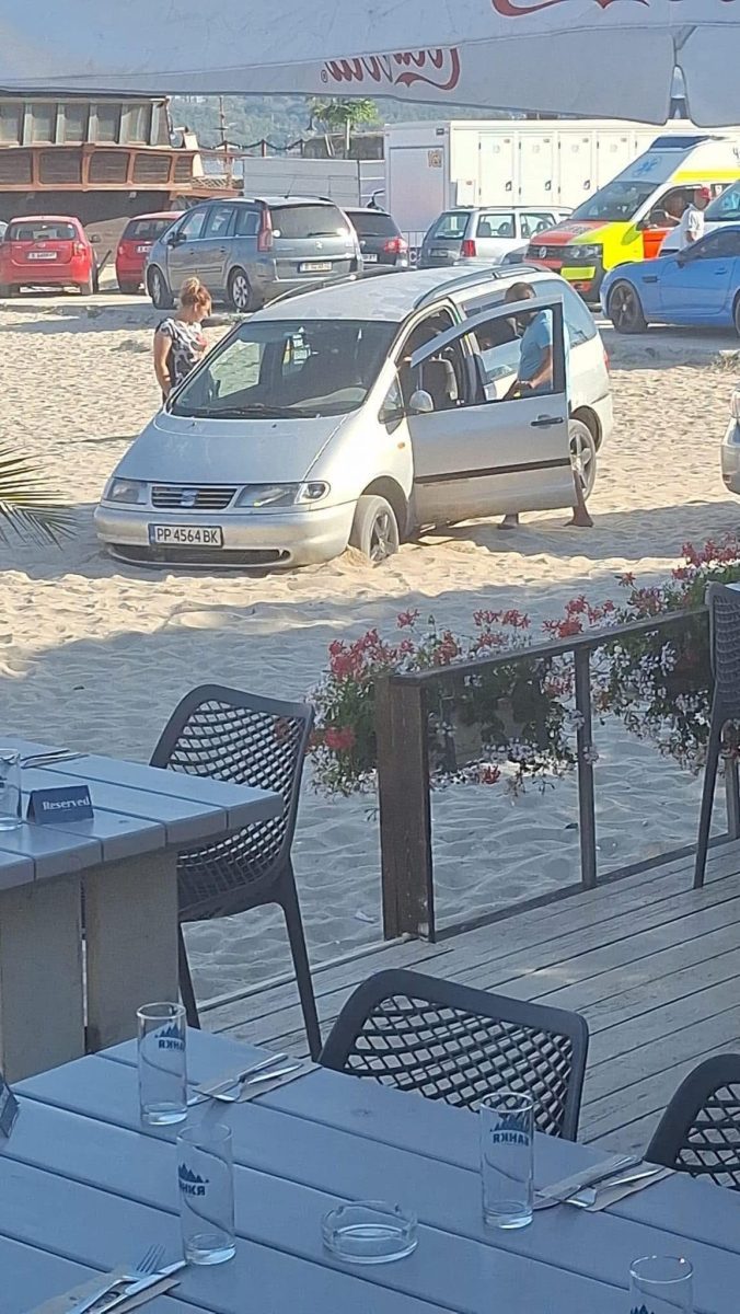 Auto zapadnuté v piesku na pláži, otvorené dvere, ľudia sa snažia vyriešiť situáciu, pozadie s terasou a kvetmi.