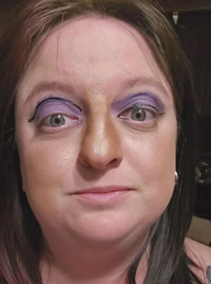 Osoba s fialovým očným tieňom a čiernou linkou, pózuje na tmavom pozadí, zvýraznený detail tváre, svetlé oči.