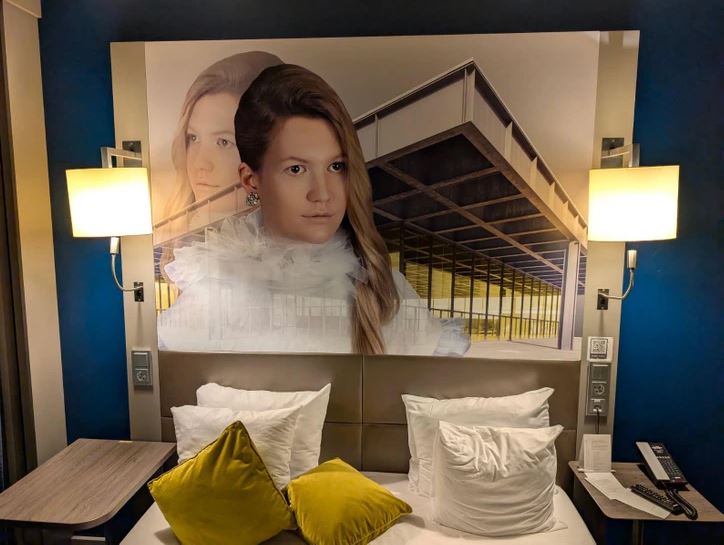 Osvetlená hotelová izba s veľkým moderným obrazom nad posteľou a vankúšmi.