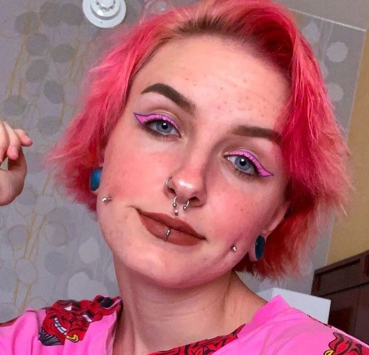 Osoba s krátkymi ružovými vlasmi a výrazným ružovým líčením očí, nosnými prsteňmi a piercings na tvári.