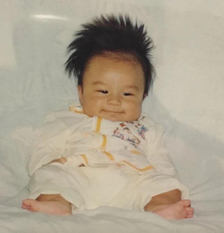 Roztomilé bábätko s roztrapatenými vlasmi v bielom oblečení sedí na mäkkej deke.