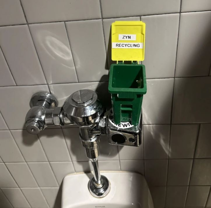 Malý žltý kontajner na recykláciu pripevnený na splachovači nad pisoárom.