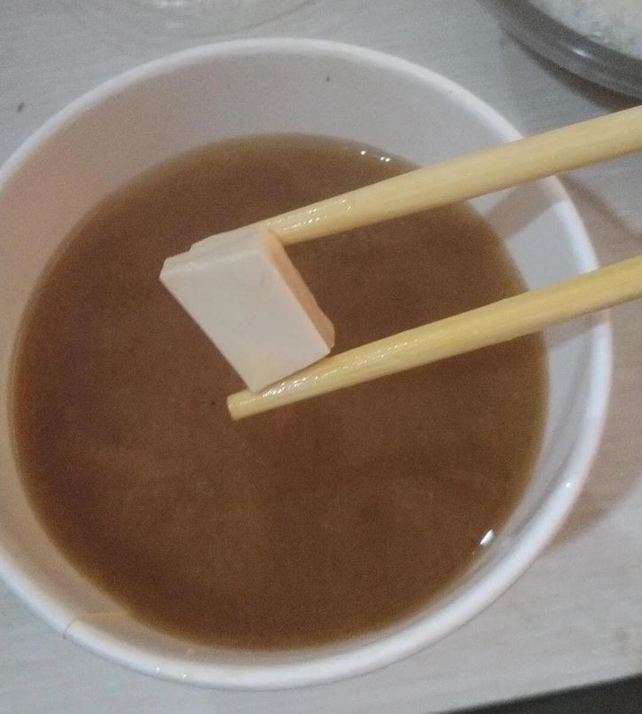 Miso polievka so štvorčekom tofu držiacim v paličkách v bielej miske na stole.