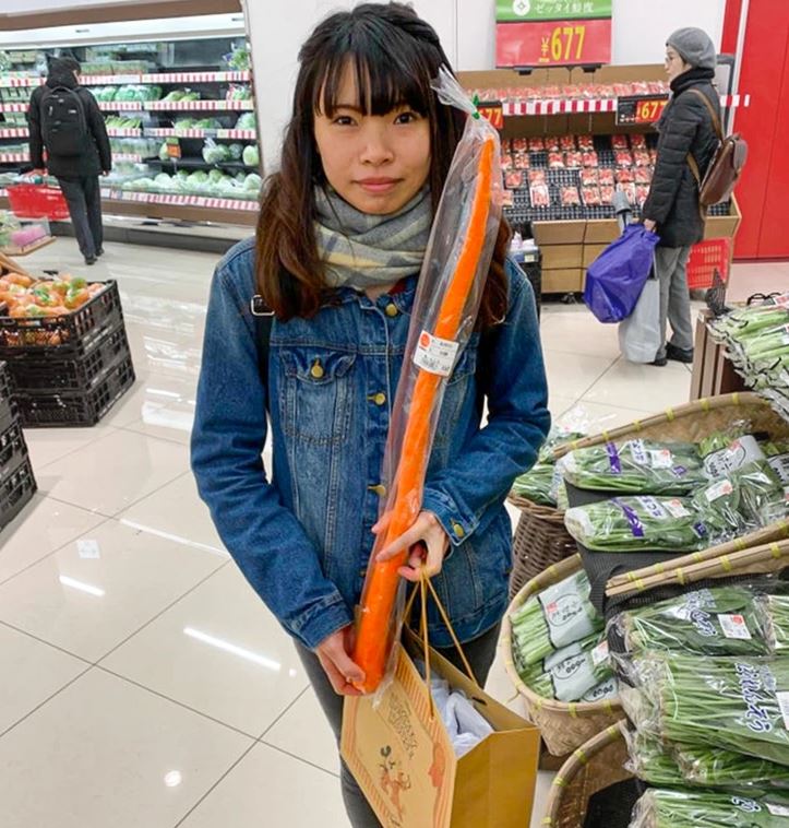 Žena v obchode drží veľkú mrkvu, okolo sú regály so zeleninou. Nakupovanie potravín, veselá nálada.