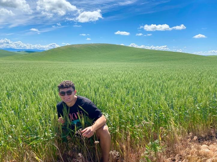 Muž v slnečných okuliaroch kľačí v zelenej pšeničnej poli pod modrou oblohou s oblakmi.