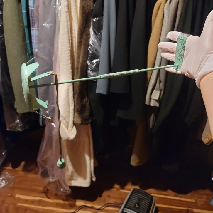 Osoba v rukavici drží zelený teleskopický nástroj v skrini plnej oblečenia.