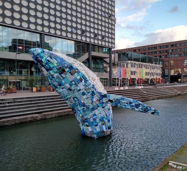 Plastová socha veľryby vyčnieva z vody pred modernou budovou v mestskom prostredí.