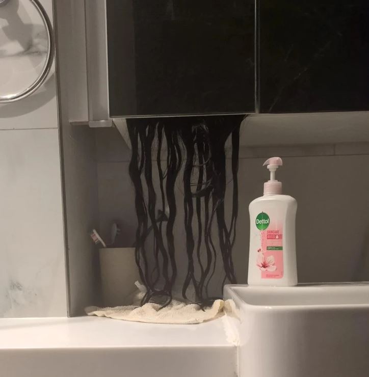 Dlhé čierne vlasy visiace zo skrinky nad umývadlom vedľa fľaše mydla Dettol v kúpeľni.