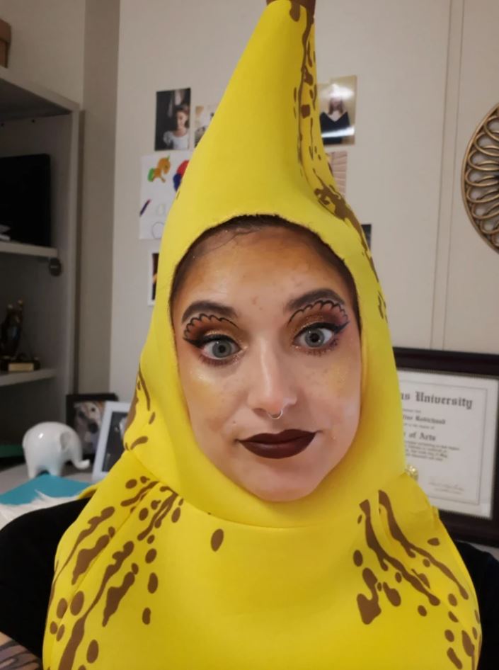 Osoba v kostýme banánu s výrazným líčením stojí v interiéri s certifikátmi na stene.