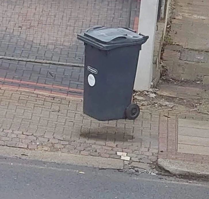 Plávajúci smetný kôš na betónovom chodníku pred garážou, vyvoláva optickú ilúziu levitácie.