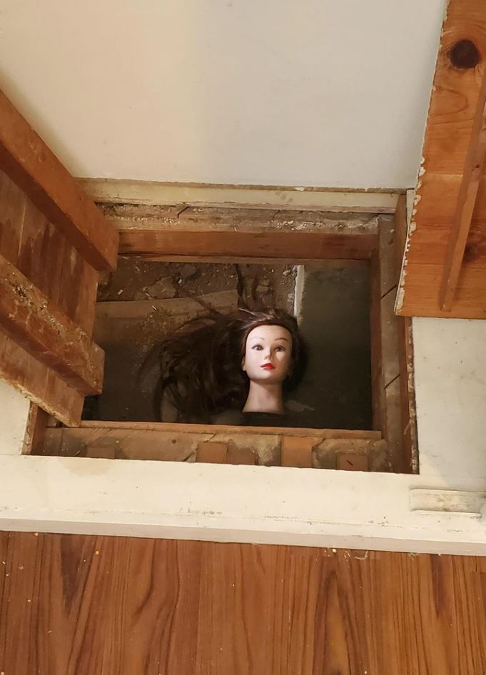 Bábková hlava zobrazujúca tmavé vlasy a je umiestnená v podlahe domu medzi drevenými trámami.