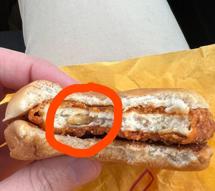 Burger s mäsovou plackou a syrom, držiaci v ruke na žltom obale, s červeným kruhom zvýrazneným detailom.