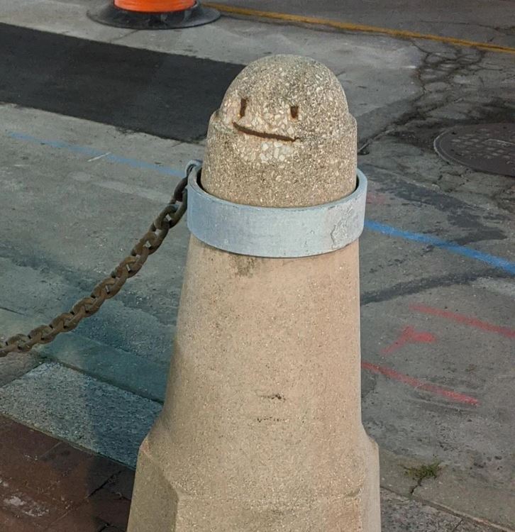 Betónový stĺpik s úsmevom na tvári, obklopený reťazou na mestskom chodníku.