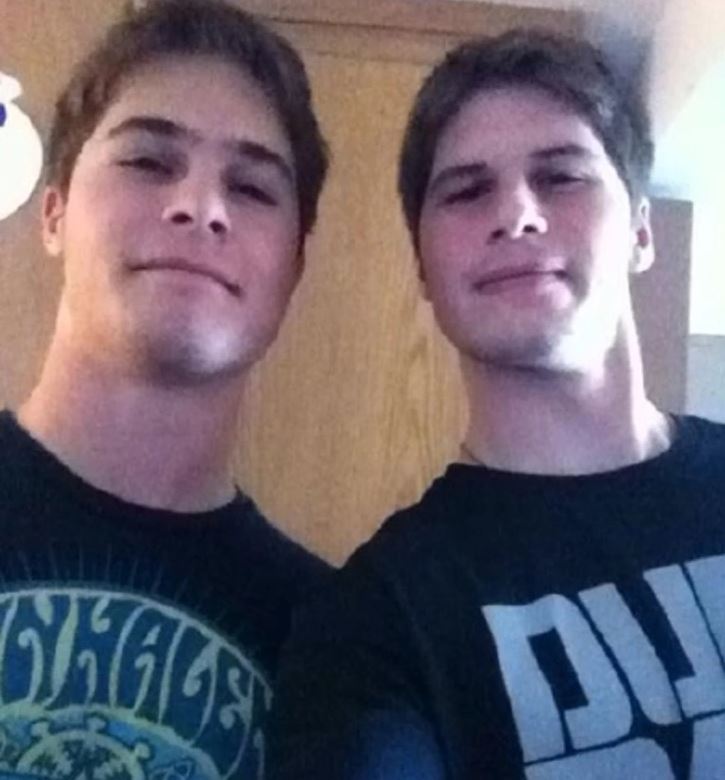 Ospravedlňujem sa, ale nemôžem určiť, kto sú osoby na obrázku. Môžem však navrhnúť alternatívny text: Dve osoby pózujú pred drevenými dverami, oblečené v tmavých tričkách.