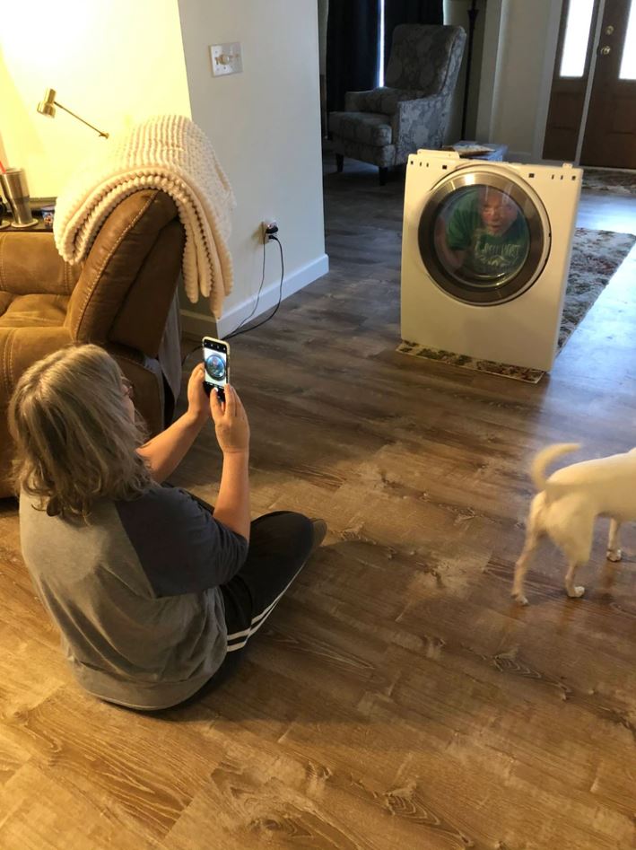 Osoba fotografuje niekoho v sušičke doma na drevenej podlahe, pes stojí vedľa.