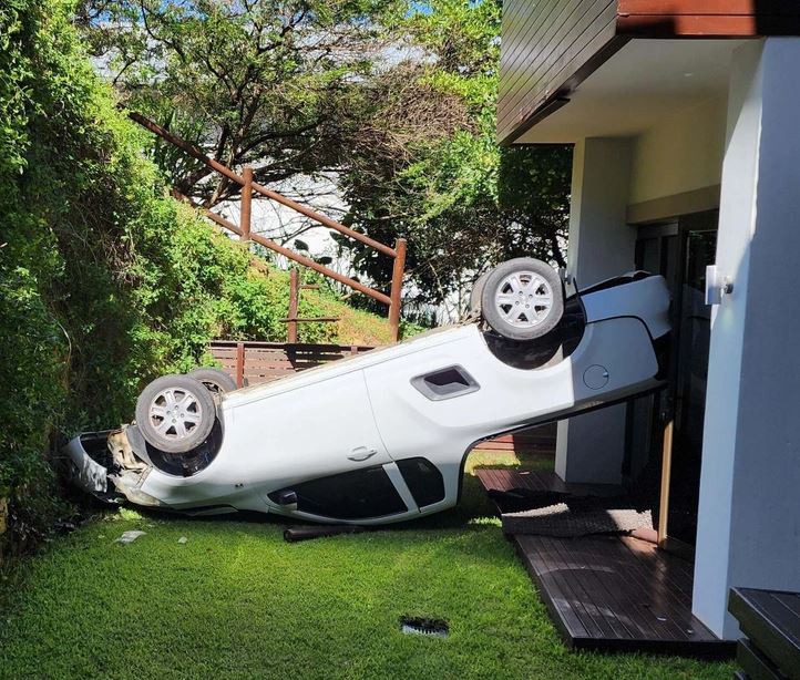 Auto prevrátené na strechu pri dome, leží na tráve medzi zeleňou, vedľa drevom pokrytej terasy.
