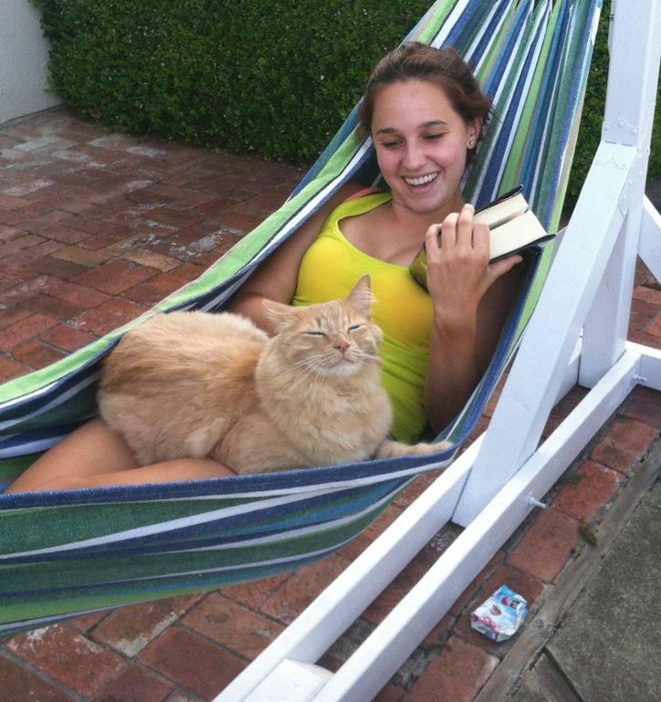 Žena relaxuje v hojdacej sieti s knihou a mačkou, užívajúc si pokojný deň vonku.
