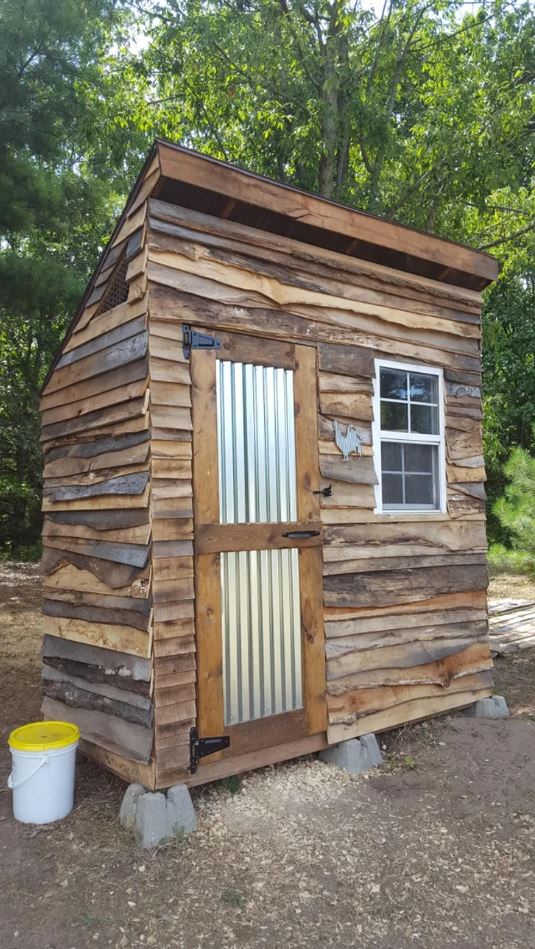 Drevená kôlňa s plechovými dverami a oknom, obklopená zeleňou.