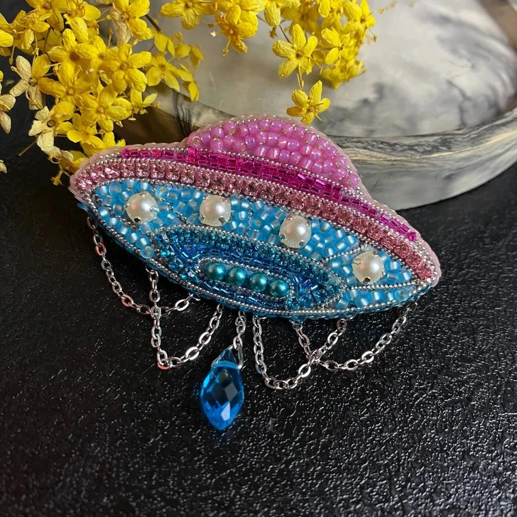 Pestrofarebná UFO brošňa zdobená korálkami a perlami, s retiazkami a modrým príveskom na čiernom pozadí.