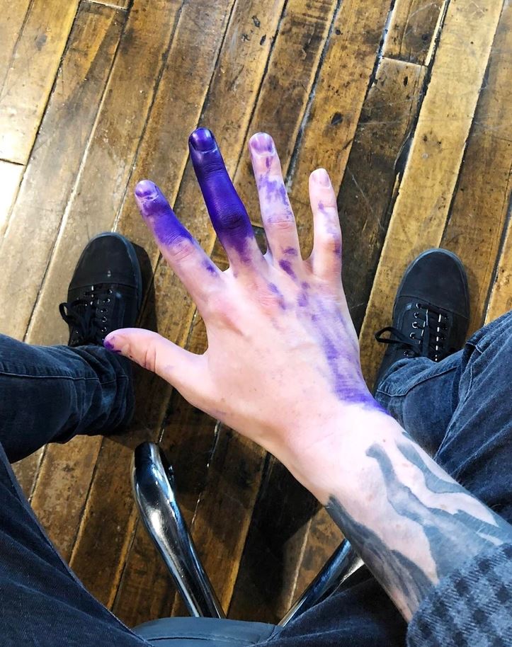 Ruka zafarbená na fialovo, sedí na drevenej podlahe v čiernych topánkach a modrých nohaviciach.