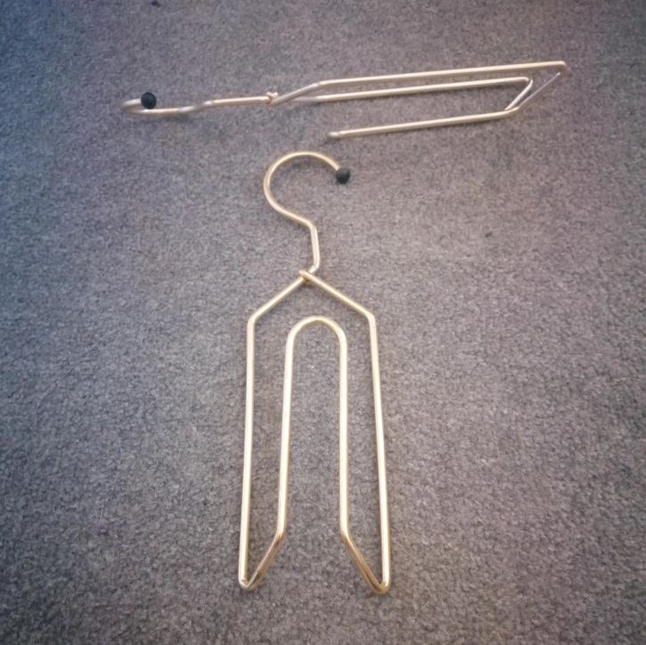 Kovový vešiak na nohavice položený na sivom koberci so zahnutým hákom a jednoduchou konštrukciou.