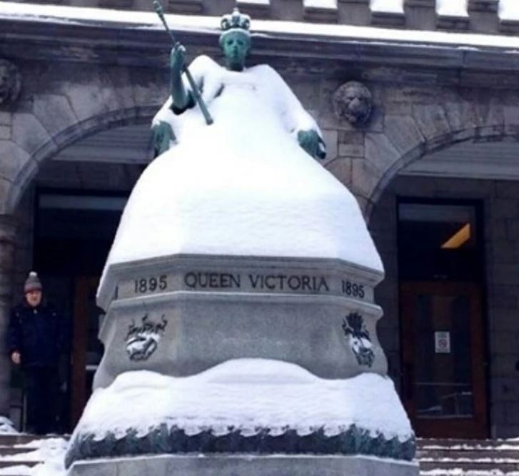 Snehom pokrytá socha kráľovnej Viktórie pred historickou budovou za zimného dňa.