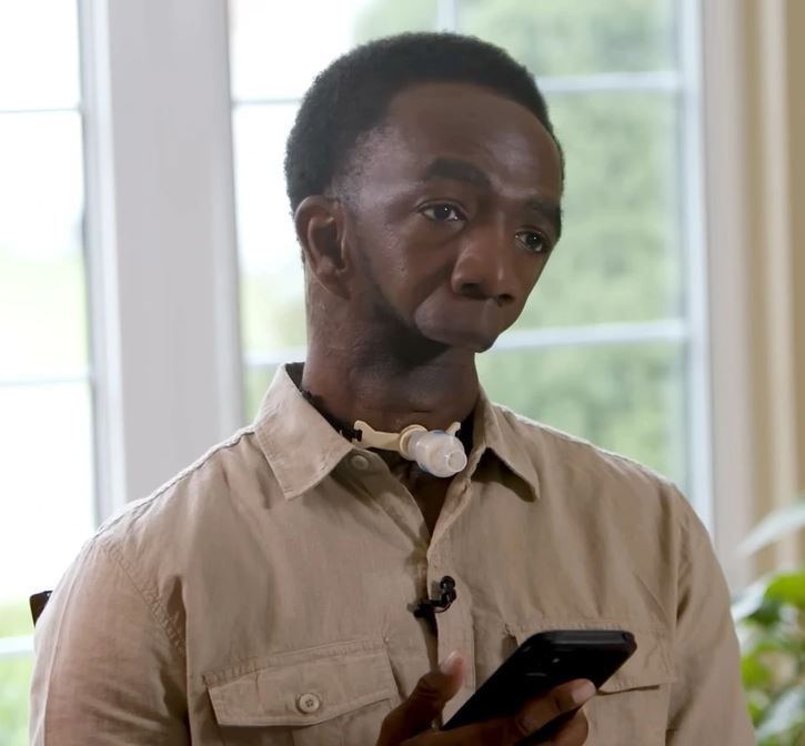 Osoba s tracheostómiou drží mobilný telefón v miestnosti s veľkými oknami v pozadí.