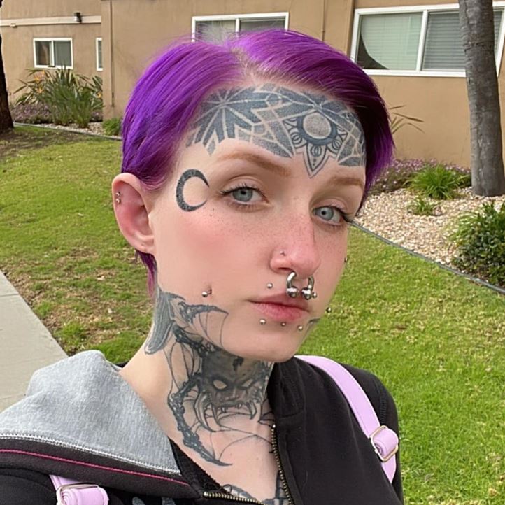 Osoba vonku s fialovými vlasmi, výraznými tetovaniami na tvári a krku a piercingmi na nos a pery.