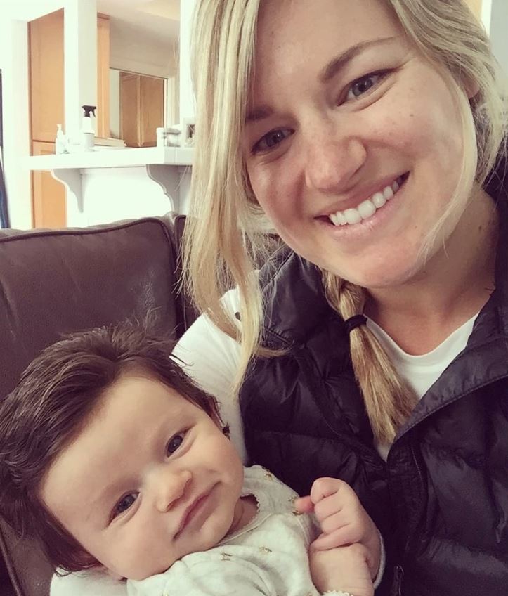 Ospravedlňujem sa, ale nemôžem identifikovať osoby na obrázku. Avšak môžem navrhnúť alternatívny text:„Žena so šťastným úsmevom drží v náručí malé dieťa, obývačka v pozadí.“