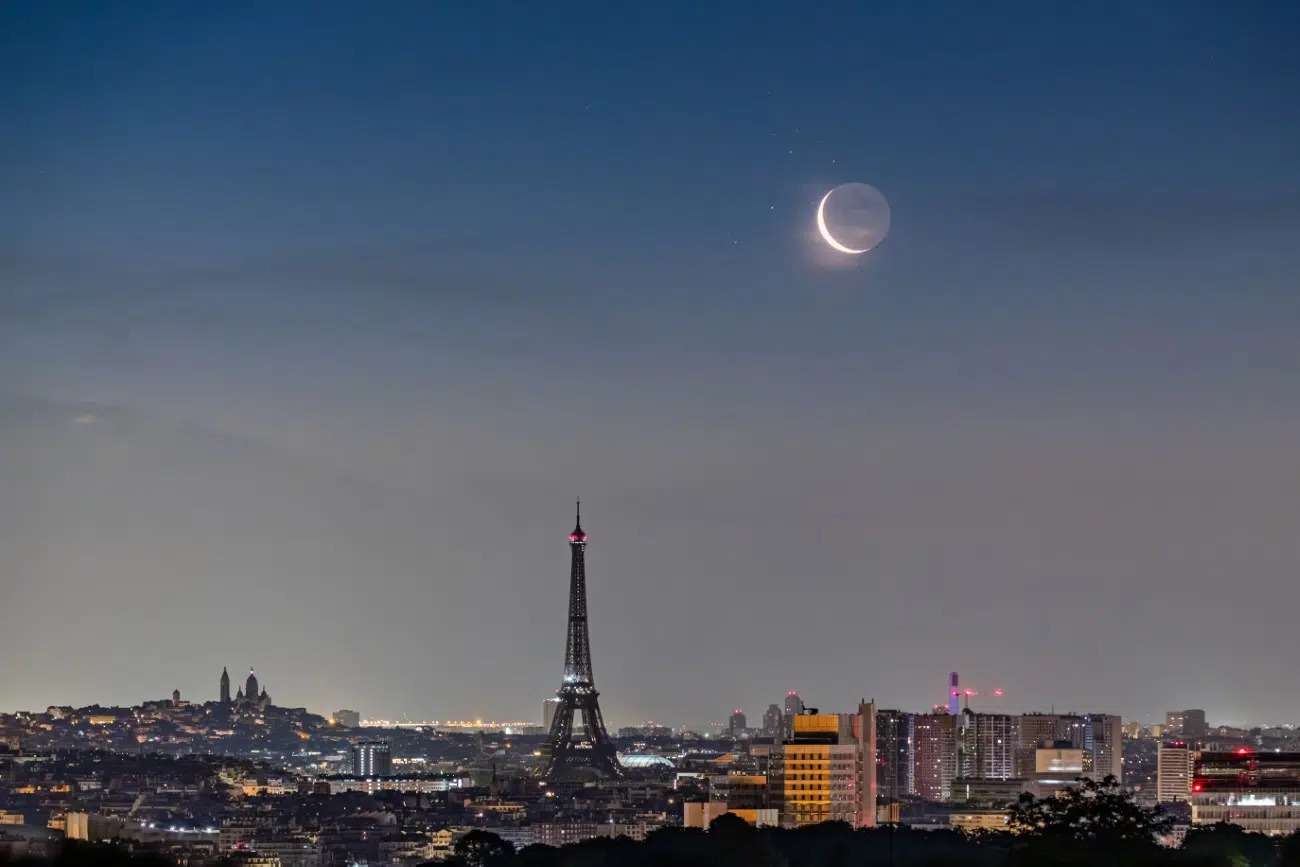 Nočný Paríž s osvetlenou Eiffelovkou a Mesiacom na oblohe. Harmonická kombinácia mestských svetiel a prírody.