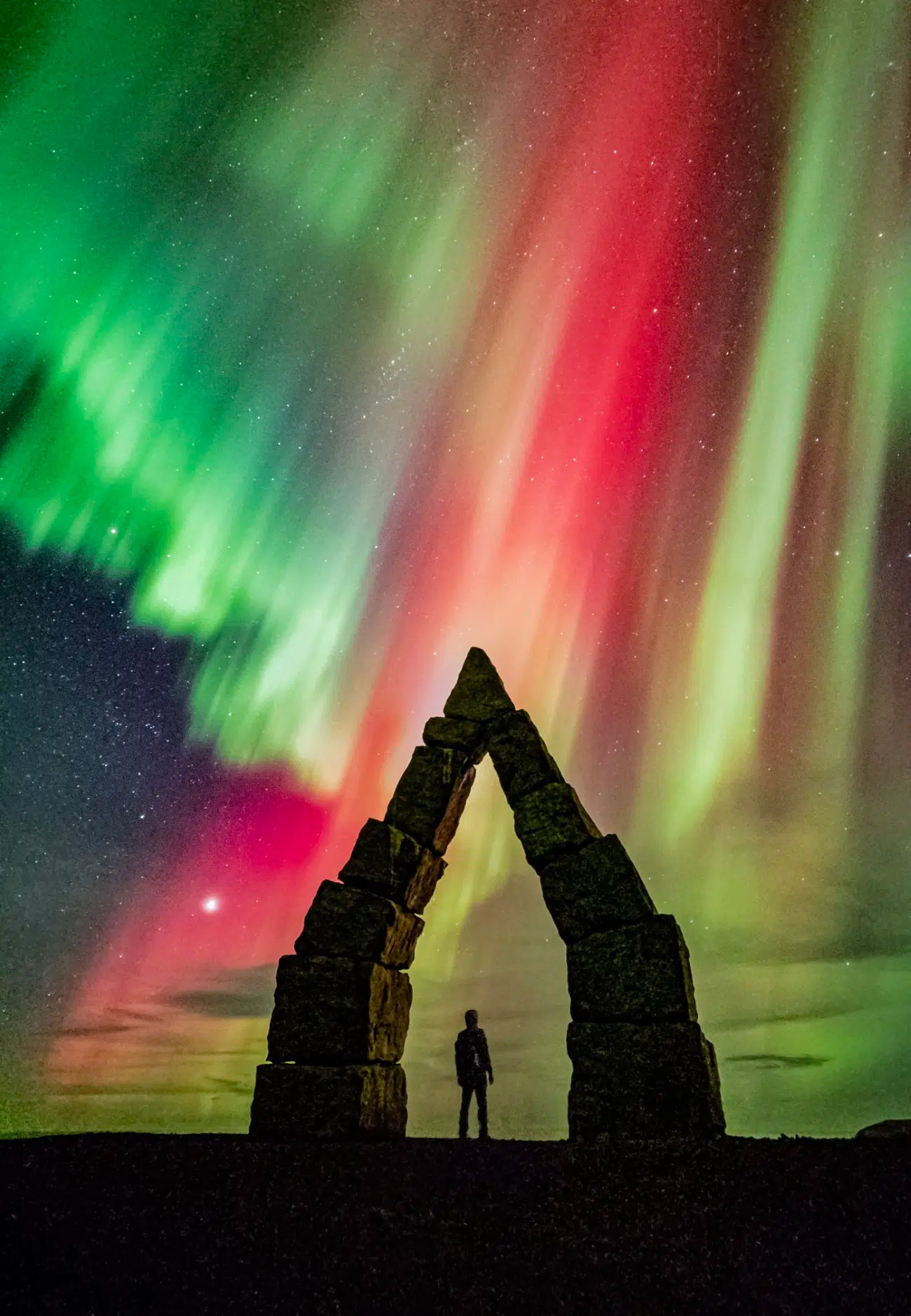 Osoba stojaca pod kamennou bránou s polárnou žiarou v pozadí v žiarivých farbách na nočnej oblohe.