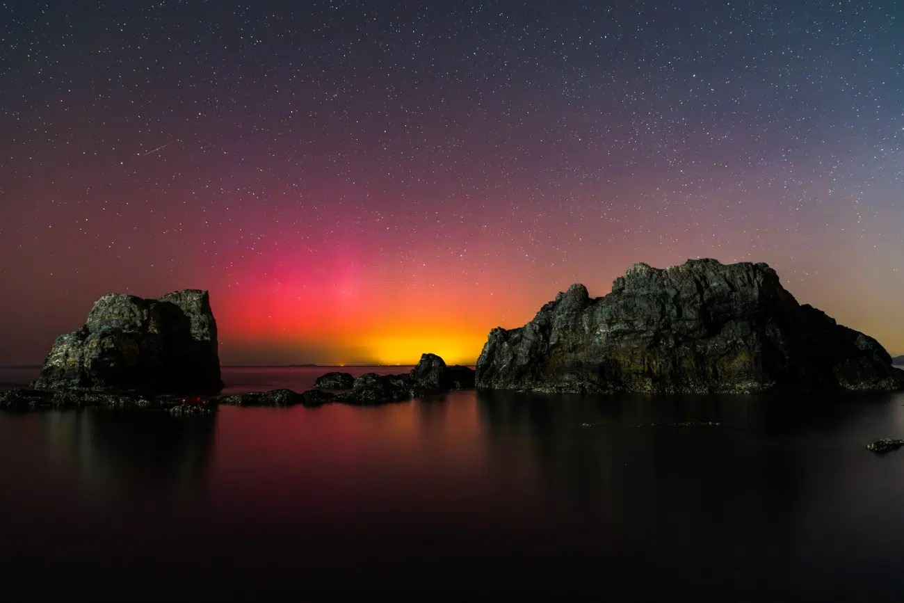 Polárna žiara nad morskou hladinou s hviezdami a dramatickými skalnatými útvarmi v popredí.